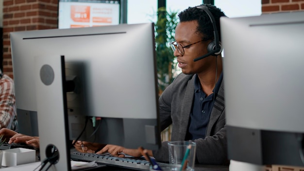 African american employee using headset at customer service job, asnwering call about telemarketing sales. Male operator working at call center office to help clients on helpline.