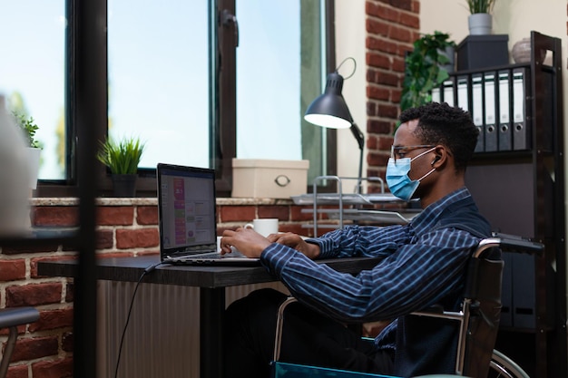 African american employee who is paralyzed wearing covid19 protection mask analyzing key performance indicators on laptop. Startup owner looking at portable computer using wireless earbuds.