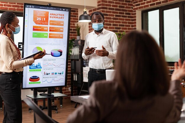 スタートアップオフィスの企業パートナーシップで働くビジネスグラフを説明する経営陣のプレゼンテーションを示すアフリカ系アメリカ人の起業家。 covid19に対する医療用フェイスマスクを備えたビジネスチーム