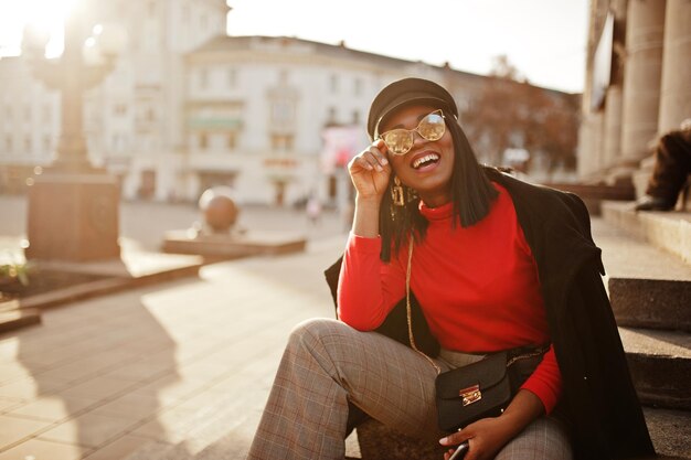 코트와 뉴스보이 모자 선글라스에 아프리카계 미국인 패션 소녀가 거리에서 포즈를 취했습니다.
