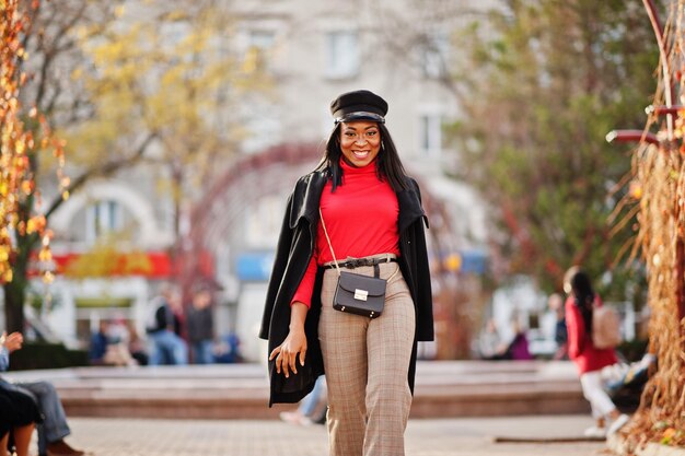 ストリートでポーズをとったハンドバッグとコートとキャスケットのアフリカ系アメリカ人のファッションの女の子