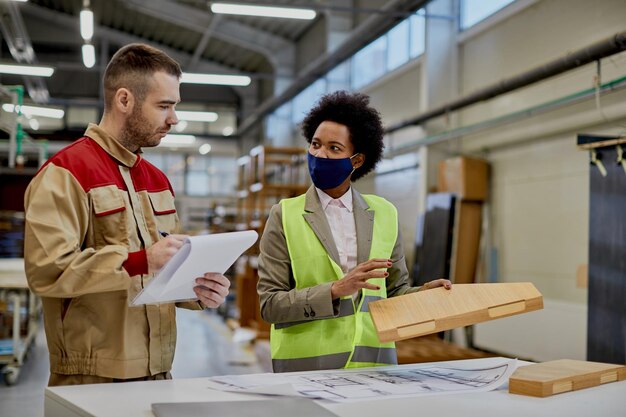 Афроамериканка-инспектор разговаривает с рабочим во время осмотра обработанной древесины на фабрике