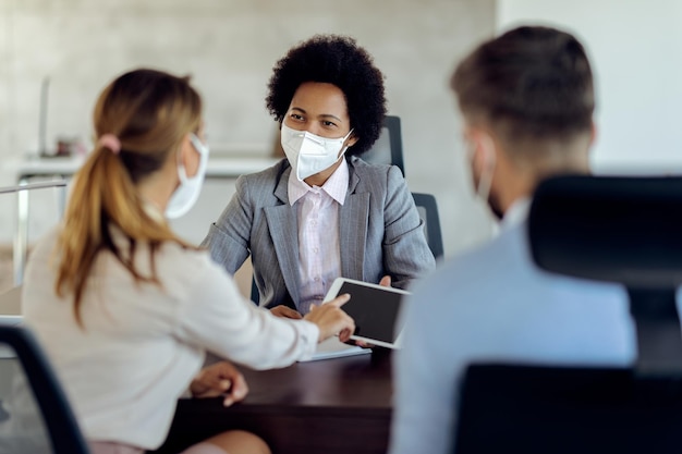 African American financial advisor and her clients using digital tablet while wearing protective face masks during the meeting