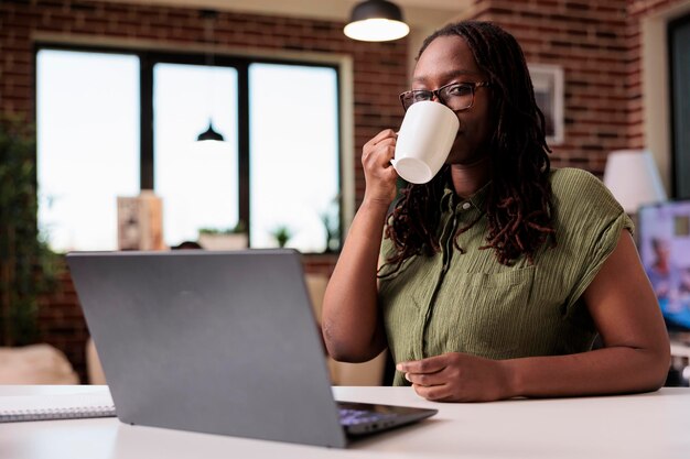 African american freelancer taking a break from remote work drinking coffee or tea at desk with laptop looking at screen. Student at home watching video content on portable computer in living room.