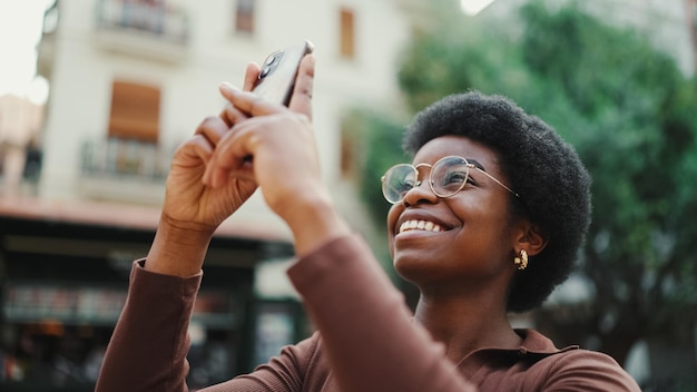African American girl taking photo of beautiful architecture on phone outdoors Cheerful woman in glasses smiling on street