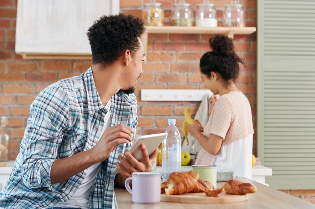 Афроамериканец смотрит на жену или девушку, просит ее дать банан, сидит за кухонным столом, пользуется современным планшетным компьютером
