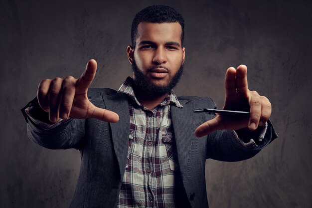 African-American guy wearing a jacket and checkered shirt is posing next to a dark textured wall.