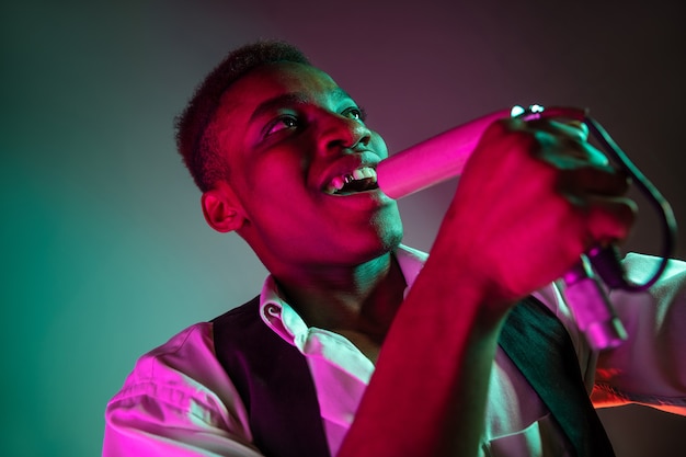 Free photo african american handsome jazz musician singing into the microphone in the studio on a neon background. music concept. young joyful attractive guy improvising. close-up retro portrait.