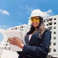 Free photo african american lady in safety helmet with notebook near building under construction