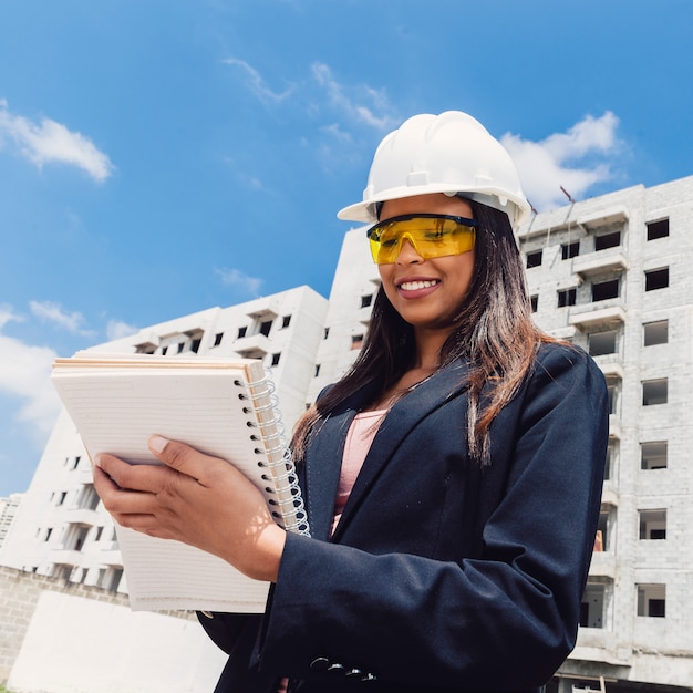 African American lady in safety helmet with notebook near building under construction