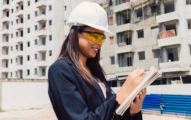 African American lady in safety helmet with notepad near building under construction