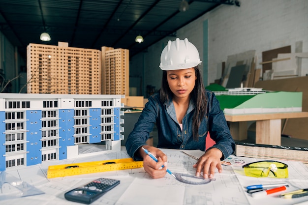 African-American lady in safety helmet working near model of building