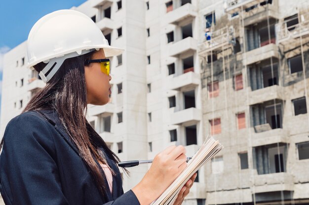 African American lady in safety helmet writing in notepad near building under construction