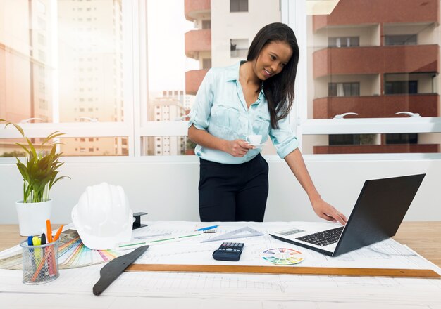 African-American lady with paper near laptop and plan on table with equipments