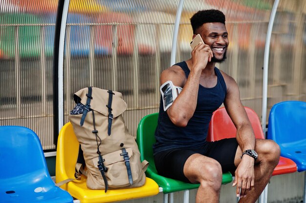 Афро-американский спортсмен в спортивной одежде с рюкзаком сидит на стуле на стадионе и разговаривает по мобильному телефону