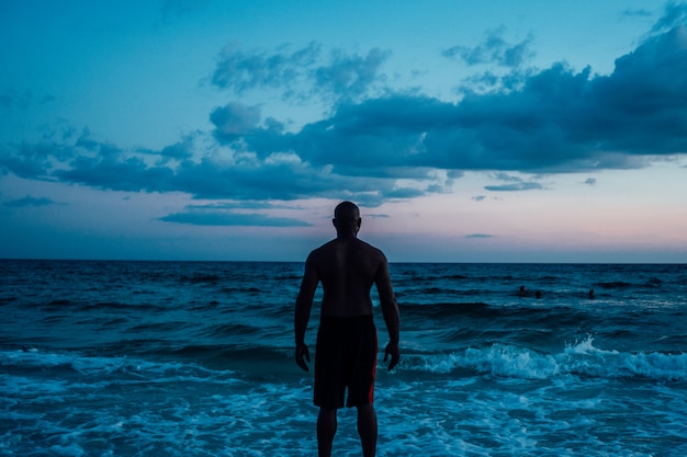 African American male standing near the sea under a blue cloudy sky shot from behind