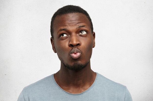 Афро-американский мужчина в повседневной серой рубашке, надувает губы и смотрит вверх с нерешительным выражением лица