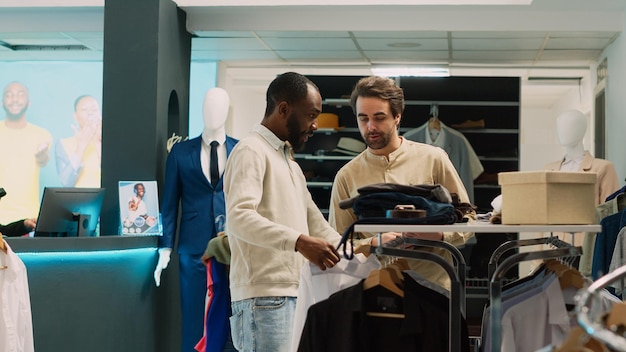 Free photo african american man asking assistant for help with shirts, looking to buy formal wear on hangers. retail store employee talking to client about shopping for fashionable clothes. tripod shot.