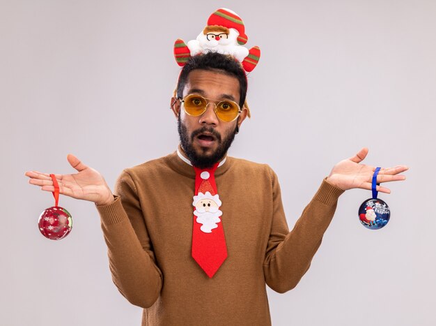 African american man in brown sweater and santa rim on head with funny red tie holding christmas balls looking at camera confused standing over white background