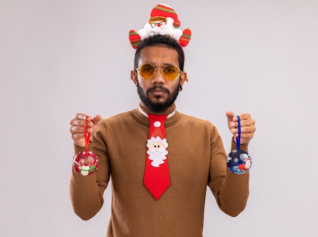 African american man in brown sweater and santa rim on head with funny red tie holding christmas balls looking at camera with sad expression standing over white background