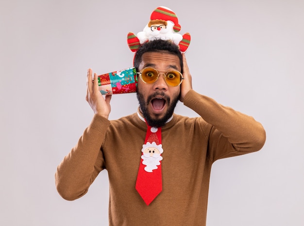 African american man in brown sweater and santa rim on head with funny red tie holding colorful paper cup over ear looking surprised standing over white background