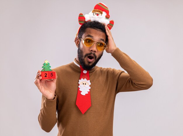 African american man in brown sweater and santa rim on head with funny red tie holding toy cubes with date twenty five looking at camera confused with hand on his head standing over white background