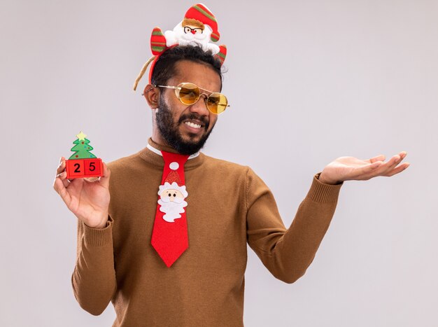 African american man in brown sweater and santa rim on head with funny red tie holding toy cubes with date twenty five looking at camera displeased with arm out standing over white background
