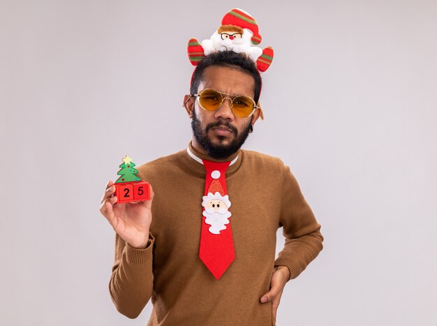 Free photo african american man in brown sweater and santa rim on head with funny red tie holding toy cubes with date twenty five looking at camera with serious face standing over white background