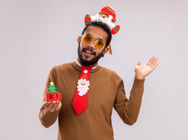 African american man in brown sweater and santa rim on head with funny red tie holding toy cubes with new year date looking at camera smiling with happy face standing over white background