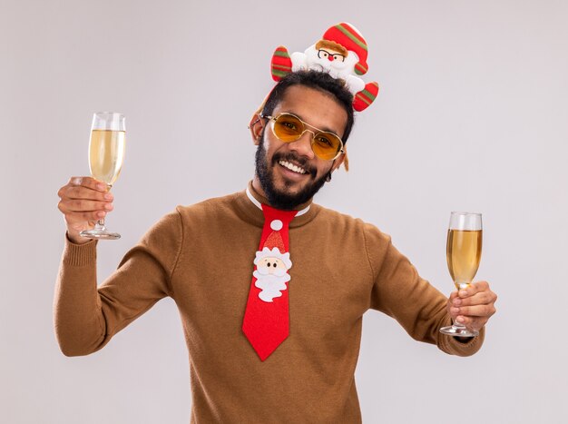 African american man in brown sweater and santa rim on head with funny red tie holding two glasses of champagne happy and cheerful smiling standing over white background