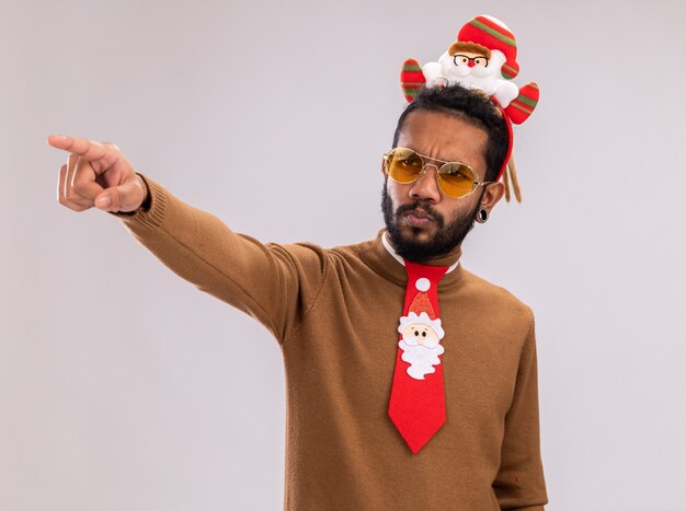 African american man in brown sweater and santa rim on head with funny red tie looking aside with serious face pointing with index finger at something standing over white background