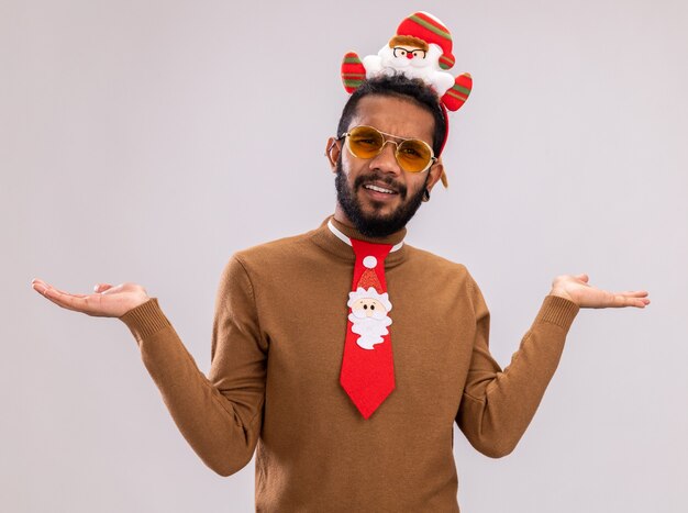 African american man in brown sweater and santa rim on head with funny red tie looking at camera confused and displeased standing over white background