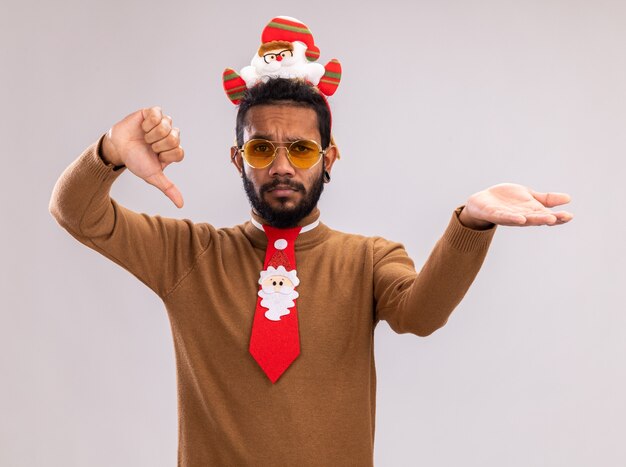 African american man in brown sweater and santa rim on head with funny red tie looking at camera showing thumbs down presenting with arm standing over white background