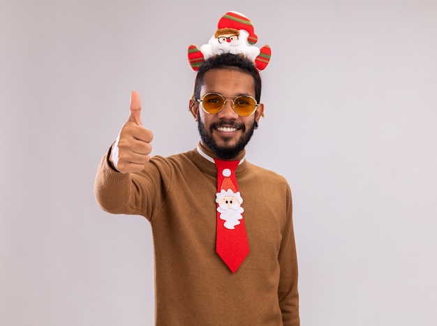 African american man in brown sweater and santa rim on head with funny red tie looking at camera smiling cheerfully showing thumbs up standing over white background