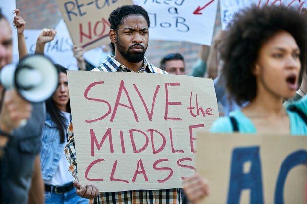 African American man carrying banner with 'save the middle class' inscription while protesting with crowd of people on city streets