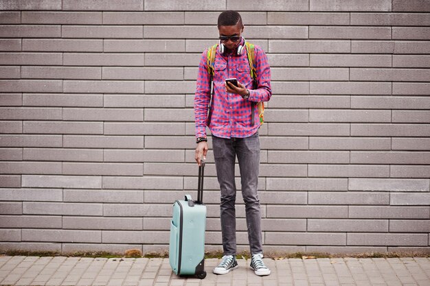 체크 무늬 셔츠 선글라스와 이어폰과 가방과 배낭을 입은 아프리카 계 미국인 남자 휴대 전화를 들고 벽에 흑인 여행자