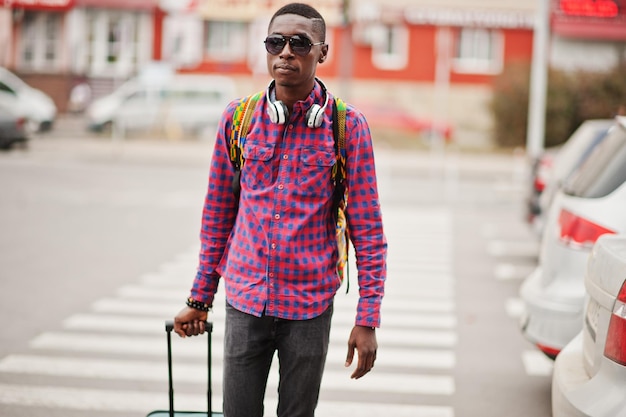 Uomo afroamericano in camicia a scacchi occhiali da sole e auricolari con valigia e zaino viaggiatore uomo nero che cammina sul passaggio pedonale