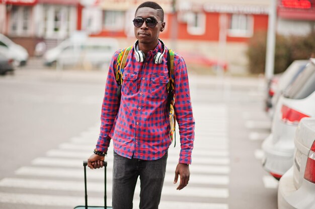 체크 무늬 셔츠 선글라스와 이어폰에 가방과 배낭을 입은 아프리카 계 미국인 남자 횡단 보도를 걷는 흑인 여행자