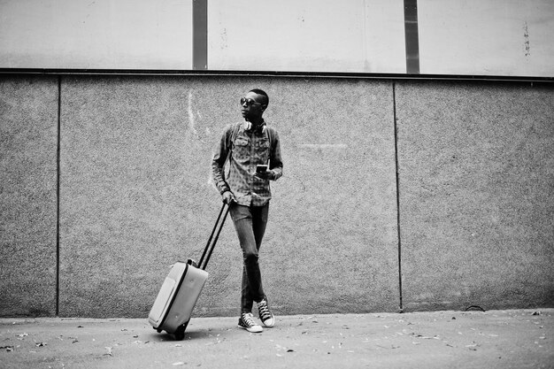 Африканский американец в солнцезащитных очках в клетчатой рубашке и наушниках с чемоданом Чернокожий путешественник у стены с мобильным телефоном