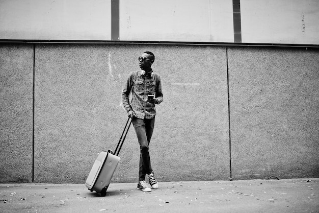 African american man in checkered shirt sunglasses and earphones with suitcase Black man traveler against wall holding mobile phone