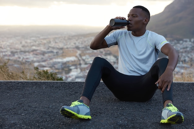 African American man drinks fresh water from bottle, rests on asphalt, sits against mountain outdoor, feels relaxed, dressed in casual t shirt, sneakes and trousers. Relaxation concept