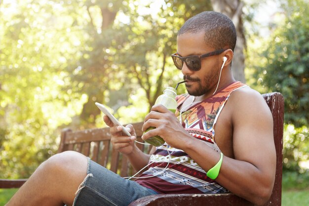 African American man in headphones sitting on bench in public park listening to songs on cell phone, checking e-mail using Internet-enabled electronic device, texting friends via social networks