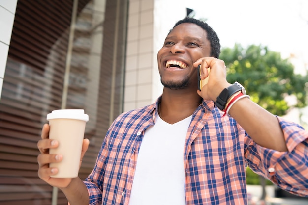 Афро-американский мужчина держит чашку кофе и разговаривает по телефону во время прогулки по улице.