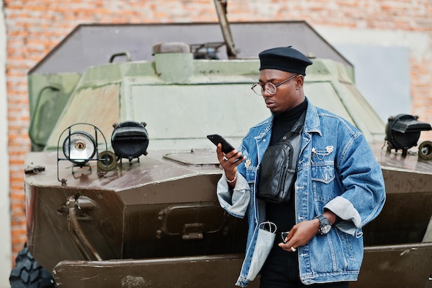 Free photo african american man in jeans jacket beret and eyeglasses speaking on phone against btr military armored vehicle