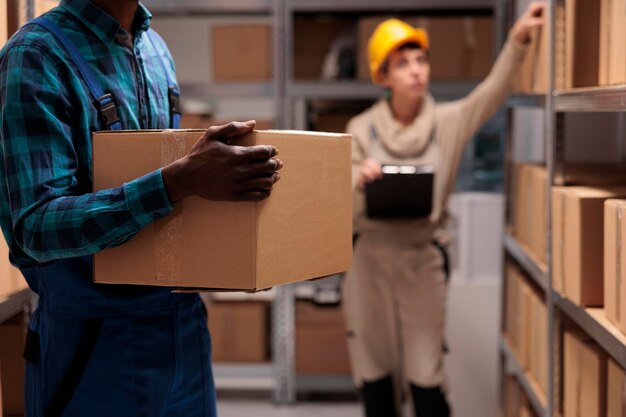 Афроамериканец поднимает тяжелую посылку на распределительном складе, несет картонную коробку на полку склада. Загрузчик склада держит коробку и работает в отделе логистики для хранения товаров