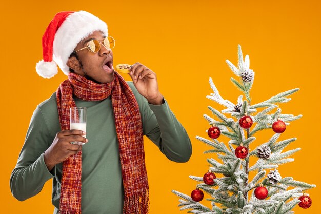 African american man in santa hat and scarf around neck eating cookie holding glass of milk standing next to a christmas tree over orange background