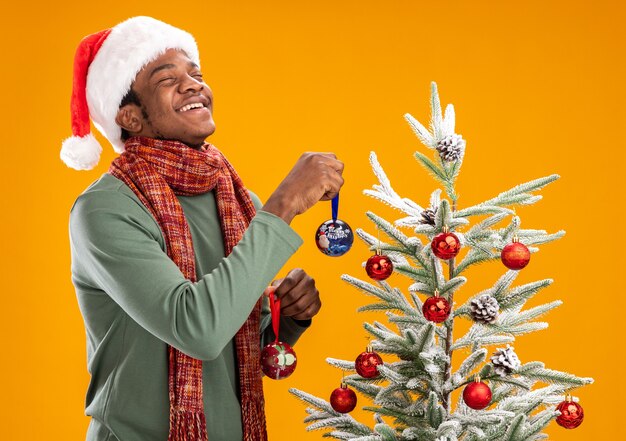 Афро-американский мужчина в новогодней шапке и шарфе на шее, висящий на елке новогодними шарами, счастливый и веселый, стоя над оранжевой стеной