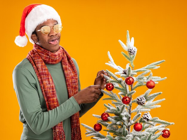 African american man in santa hat and scarf around neck hanging christmas balls on a christmas tree happy and positive standing over orange background
