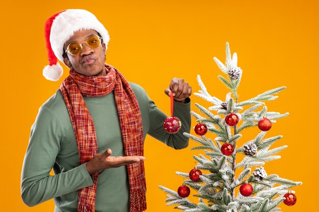 オレンジ色の背景の上のクリスマスツリーの横に立って不機嫌そうに見えるボールを保持している首の周りのサンタの帽子とスカーフのアフリカ系アメリカ人の男