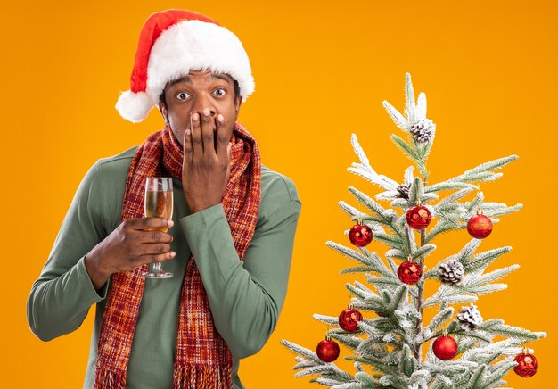 African american man in santa hat and scarf around neck holding glass of champagne looking at camera shocked covering mouth with hand next to christmas tree over orange background